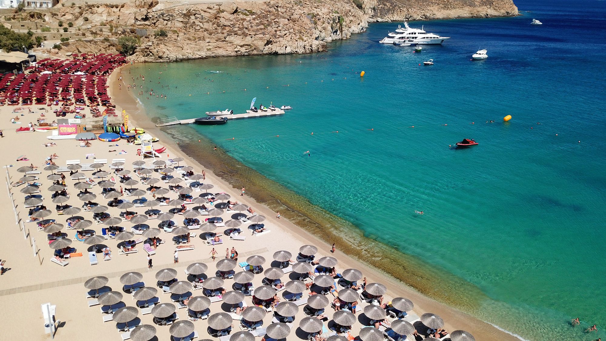 A sandy beach on the south coast of Mykonos with sunbeds, turquoise water and yachts