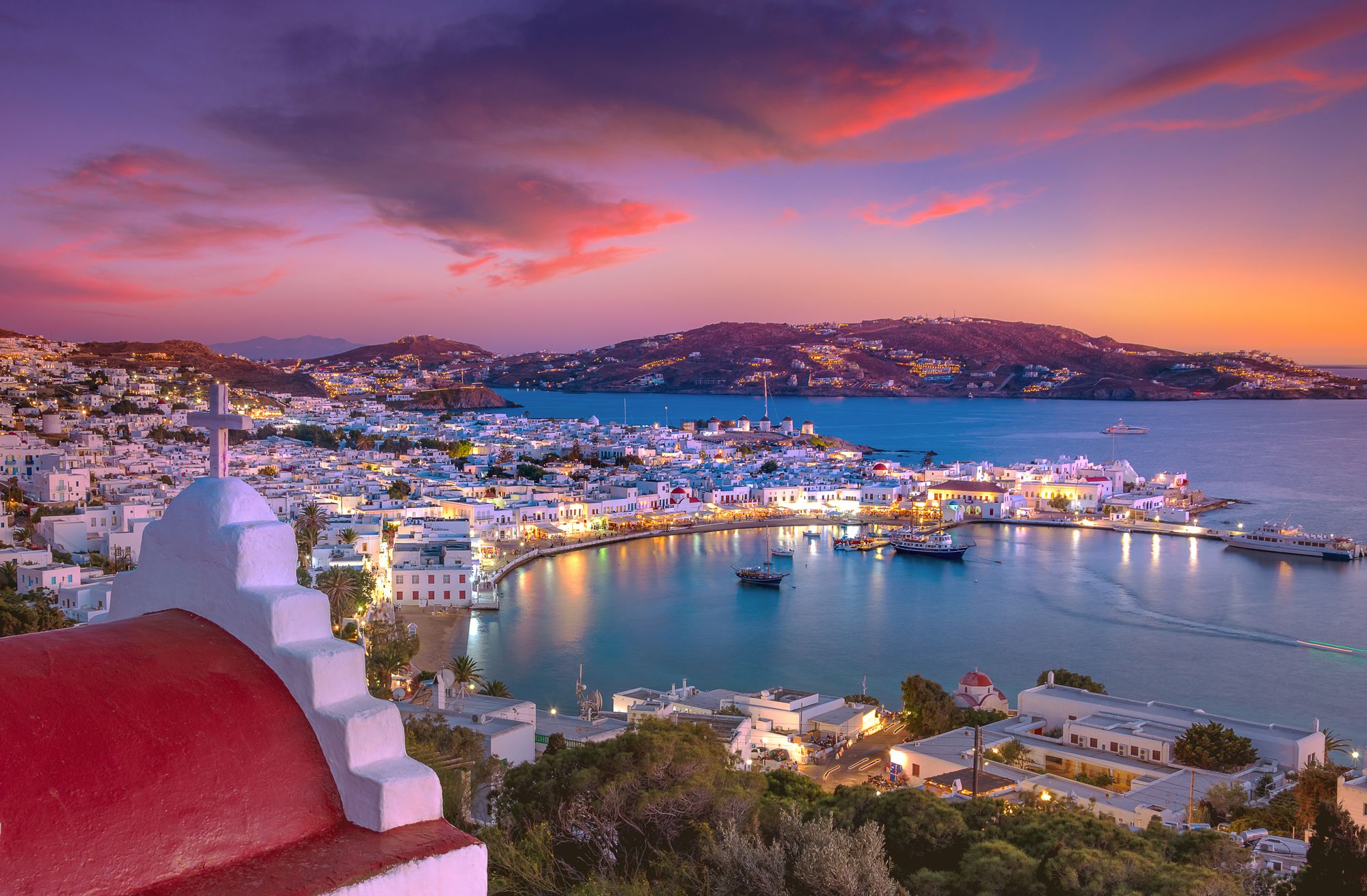 Mykonos Town and the old port lit up at dusk, seen past a chapel with a red roof